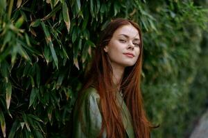 un hermosa joven mujer con volador rojo pelo camina en el ciudad en el parque en contra un antecedentes de verde bambú hojas, primavera en el ciudad foto