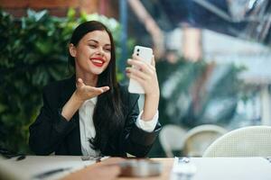 elegante mujer blogger turista se sienta en un café con un teléfono en su manos lee un mensaje, móvil comunicación y Internet en un viaje, vídeo llamar, Lanza libre trabajo en línea, sonrisa con dientes foto