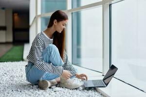 A woman smiling with teeth sits with a laptop by the large windows of a building in the city talking via video link through a computer, the concept of work and leisure in the big city photo