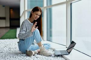 A woman smiling with teeth sits with a laptop by the large windows of a building in the city talking via video link through a computer, the concept of work and leisure in the big city photo