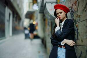 Moda mujer retrato caminando turista en elegante ropa con rojo labios caminando abajo un estrecho ciudad calle, viajar, cinematográfico color, retro Clásico estilo, dramático en contra un pared con pintada. foto