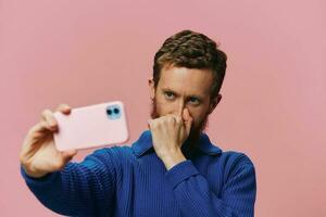 Portrait of a redheaded man with phone in hand taking selfies and photos on his phone with a smile on a pink background, blogger