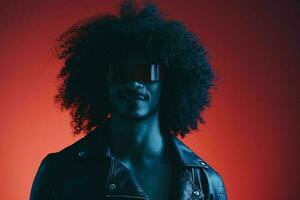 Portrait of fashion man with curly hair on red background with stylish glasses, multicultural, colored light, black leather jacket trend, modern concept. photo