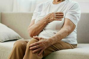 Elderly woman severe chest pain sitting on the sofa, health problems in old age, poor quality of life. Grandmother with gray hair holds her chest with her hands, women's health, breast cancer. photo