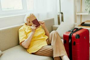 contento mayor mujer con pasaporte y viaje boleto lleno un rojo maleta, vacaciones y salud cuidado. sonriente antiguo mujer felizmente se sienta en el sofá antes de el viaje. foto