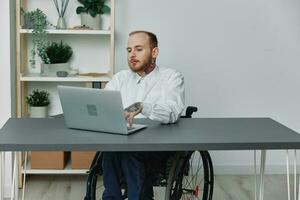 un hombre en un silla de ruedas un empresario con tatuajes en el oficina trabajos a un computadora portátil, integración dentro sociedad, el concepto de trabajando un persona con discapacidades, libertad desde social marcos foto