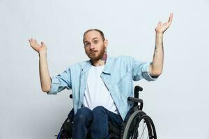 A man in a wheelchair and pointing a finger at, copy space, with tattoos on his hands sits on a gray studio background, health concept a person with disabilities, a real person photo