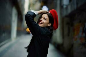 Fashion woman smile with teeth and fun tourist in stylish clothes in jacket and red beret walking down narrow city street flying hair, travel, French style, cinematic color, retro vintage style. photo