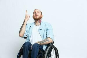 A man in a wheelchair and pointing a finger at, copy space, with tattoos on his hands sits on a gray studio background, health concept a person with disabilities, a real person photo