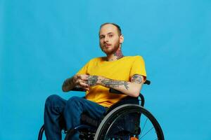un hombre en un silla de ruedas problemas con el musculoesquelético sistema mira a el cámara en un camiseta con tatuajes en su brazos se sienta en un azul estudio fondo, lleno vida, real persona, salud concepto foto