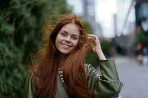 Portrait of a beautiful young woman in the city looking into the camera smile with teeth with red flying hair in fashionable clothes in the city against of bamboo in spring, lifestyle in the city photo