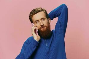 Portrait of a man with a phone in his hands doing looking at it and talking on the phone, on a pink background. Communicating online social media, lifestyle photo