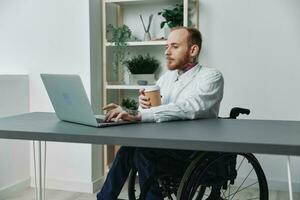 un hombre en un silla de ruedas un empresario con tatuajes en el oficina trabajos a un ordenador portátil con café, integración dentro sociedad, el concepto de trabajando un persona con discapacidades, libertad desde social marco de referencia foto