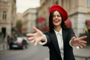 Moda mujer sonrisa con dientes en pie en el calle en frente de el ciudad turista seguir yo elegante ropa con rojo labios y rojo boina, viajar, cinematográfico color, retro Clásico estilo, urbano moda. foto