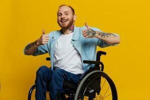 un hombre en un silla de ruedas sonrisa y felicidad, pulgar arriba, con tatuajes en su manos se sienta en un amarillo estudio fondo, el concepto de salud un persona con discapacidades foto