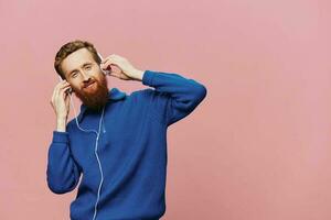 Portrait of a redheaded man wearing headphones smiling and dancing, listening to music on a pink background. A hipster with a beard. photo