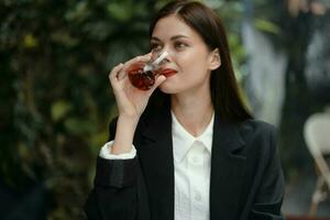 Business brunette woman with red lips smile with teeth drinks tea in a cafe from a Turkish glass mug in a white shirt and black fashion jacket on the street, summer travel, vacation in the city photo