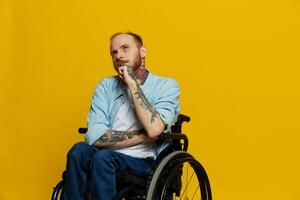 A man in a wheelchair problems with the musculoskeletal system looks at the camera thoughtfully with tattoos on his arms sits on a yellow studio background, health concept a person with disabilities photo