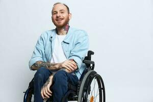 un hombre en un silla de ruedas sonrisa, Copiar espacio, con tatuajes en su brazos se sienta en un gris estudio fondo, el concepto de salud es un persona con discapacidades, un real persona foto