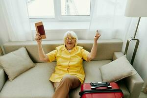 Happy senior woman with passport and travel ticket packed a red suitcase, vacation and health care. Smiling old woman joyfully sitting on the sofa before the trip raised her hands up in joy. photo