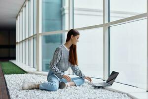 A woman freelancer sits in the office lobby by the big windows and works on her laptop online via the Internet, freelance work anywhere in the world photo