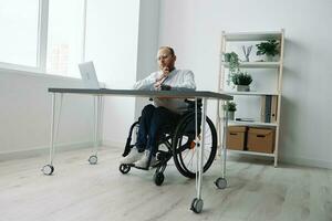 un hombre en un silla de ruedas, un empresario en el oficina trabajos a un computadora portátil, escritura abajo un plan en un computadora portátil, consideración, integración dentro sociedad, el concepto de trabajando un persona con discapacidades foto