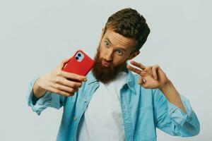 Man hipster with phone in hand blogger smile on gray background in blue shirt and white t-shirt, talking on the phone and selfies photo