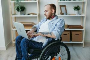 un hombre en un silla de ruedas persona de libre dedicación trabajos a un ordenador portátil a hogar, trabajo en línea, social redes y puesta en marcha, Copiar espacio, integración dentro sociedad, concepto de salud discapacitado persona, real persona de cerca foto