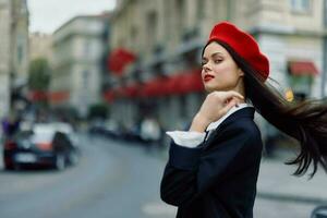 Moda mujer retrato belleza en pie en el calle en frente de el ciudad en elegante ropa con rojo labios y rojo boina, viajar, cinematográfico color, retro Clásico estilo, urbano Moda estilo de vida. foto