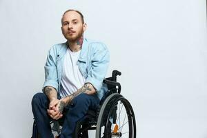 un hombre en un silla de ruedas mira pensativamente a el cámara, Copiar espacio, con tatuajes en su brazos se sienta en un gris estudio fondo, el concepto de salud un persona con discapacidades, un real persona foto