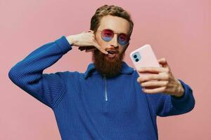 Portrait of a redheaded man with phone in hand taking selfies and photos on his phone with a smile on a pink background, blogger