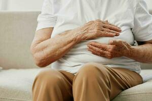 Elderly woman severe chest pain sitting on the sofa, health problems in old age, poor quality of life. Grandmother with gray hair holds her chest with her hands, women's health, breast cancer. photo