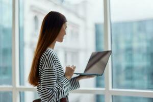 Woman with a laptop standing at the window in the office and looking out the window, work in an informal setting, business woman photo