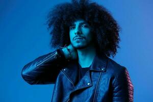 Portrait of fashion man with curly hair on blue background multinational, colored light, black leather jacket trend, modern concept. photo