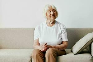 Elderly woman sits on sofa at home, bright spacious interior in old age smile, lifestyle. Grandmother with gray hair in a white T-shirt and beige trousers. photo