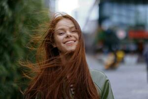 retrato de un hermosa joven niña en el ciudad mirando dentro el cámara sonrisa con dientes con rojo volador pelo en un impermeable en el ciudad en contra un antecedentes de bambú en primavera, estilo de vida en el ciudad foto