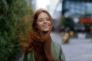 Portrait of a beautiful young girl in the city looking into the camera smile with teeth with red flying hair in a raincoat in the city against a background of bamboo in spring, lifestyle in the city photo