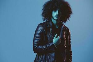 Portrait of fashion man with curly hair on blue background multinational, colored light, black leather jacket trend, modern concept. photo