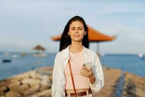 Woman smile blogger freelancer with phone in hand on vacation walking on the beach by the ocean in Bali sunset, happy travel and vacation, mobile communication, internet online photo