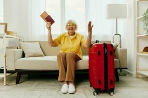 elderly woman with passport and travel ticket packed a red suitcase, vacation and health care. Smiling old woman joyfully sitting on the sofa before the trip raised her hands up the joy of retirement. photo
