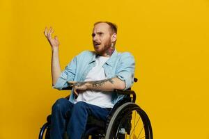 A man in a wheelchair problems with the musculoskeletal system looks at the camera is not satisfied, with tattoos on his arms sits on a yellow studio background, health concept man with disabilities photo