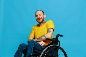 un hombre en un silla de ruedas sonrisa mira a el cámara en un camiseta con tatuajes en su brazos se sienta en un azul estudio fondo, un lleno vida, un real persona foto