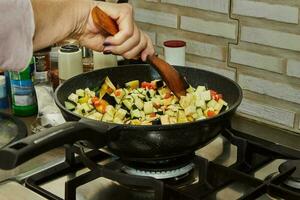 Chef stirs chopped tomatoes and eggplant into pot with ingredients on gas stove photo