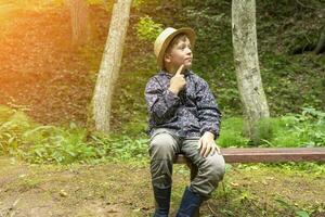 funny Caucasian boy in a traveler's hat and windbreaker sits on a bench photo