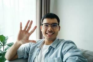 Asian man using smartphone for online video conference call with friends waving hand making hello gesture photo