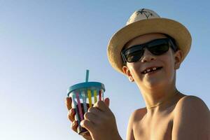 happy boy in hat and sunglasses drinking juice on the beach photo