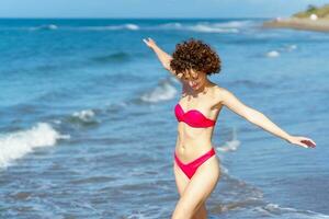 alegre joven dama en trajes de baño caminando en mar con extendido brazos foto