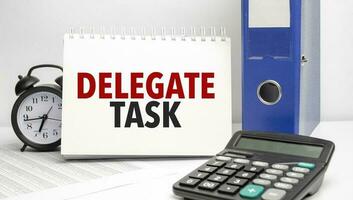 delegate task words on white notebook and calculator, black vintage alarm clock and blue paper folder photo