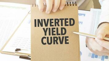 Text INVERTED YIELD CURVE on brown paper notepad in businessman hands on the table with diagram. Business concept photo