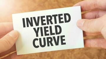 INVERTED YIELD CURVE word inscription on white card paper sheet in hands of a businessman. recap concept. red and white paper photo